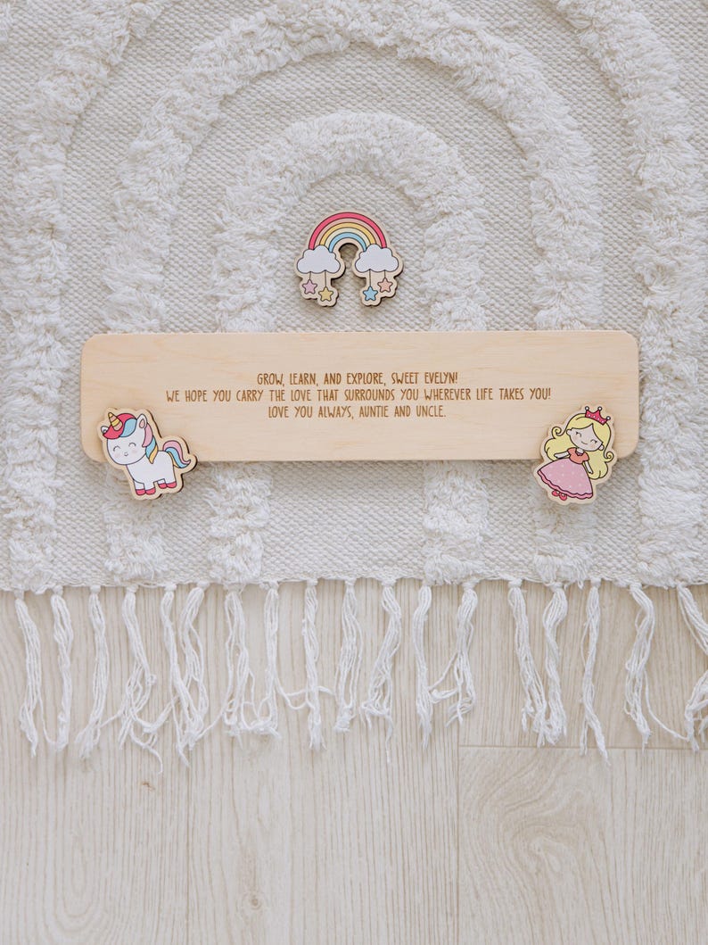 Pu&ograve; includere: Cartello in legno con la scritta "GROW, LEARN, AND EXPLORE, SWEET EVELYN!" e un arcobaleno, un unicorno e un design a principessa. Il cartello &egrave; circondato da un tappeto bianco con frange.