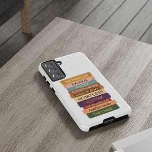 Pu&ograve; includere: Custodia bianca per telefono con una pila di dorsi di libri colorati. Ogni dorso ha un titolo, tra cui "THE FINISH LINE", "EXODUS" e "FLOCK". La custodia &egrave; su una superficie di legno marrone chiaro.
