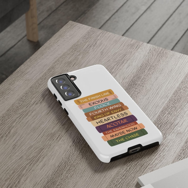 Pu&ograve; includere: Custodia bianca per telefono con una pila di dorsi di libri colorati. Ogni dorso ha un titolo, tra cui "THE FINISH LINE", "EXODUS" e "FLOCK". La custodia &egrave; su una superficie di legno marrone chiaro.