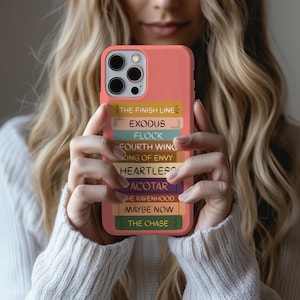 Pu&ograve; includere: Custodia per telefono color corallo con una pila di dorsi di libri colorati. I dorsi mostrano titoli come "THE FINISH LINE", "EXODUS" e "FLOCK". La custodia &egrave; tenuta da una persona con un maglione bianco.