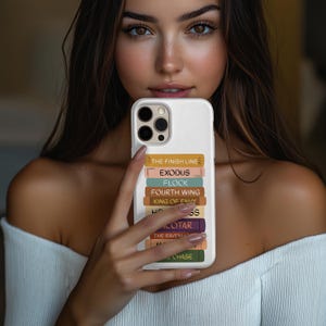 Pu&ograve; includere: Custodia bianca per telefono con una pila di dorsi di libri colorati. I titoli includono "THE FINISH LINE", "EXODUS" e "FOURTH WING". La custodia ha un ritaglio per una fotocamera a tre lenti.