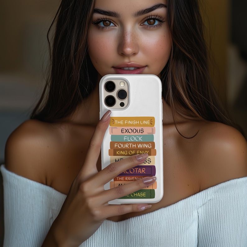 Pu&ograve; includere: Custodia bianca per telefono con una pila di dorsi di libri colorati. I titoli includono "THE FINISH LINE", "EXODUS" e "FOURTH WING". La custodia ha un ritaglio per una fotocamera a tre lenti.