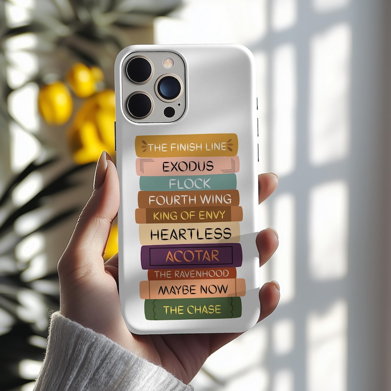 Pu&ograve; includere: Custodia per telefono bianca con un design di dorsi di libri impilati in vari colori. I dorsi mostrano titoli come "THE FINISH LINE", "EXODUS" e "ACOTAR". La custodia ha una finitura lucida e un ritaglio per la fotocamera.
