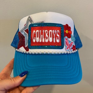 May include: A blue and white trucker hat with a red, white, and blue star, a red flower, and a butterfly patch. The hat has a white brim with a pearl trim and a red patch that says "COWBOYS" in white letters.