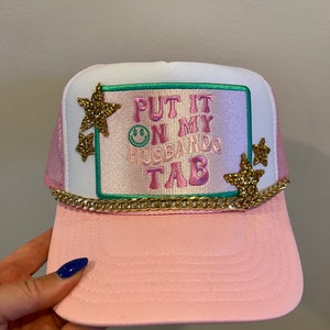 May include: Pink and white trucker hat with a green and pink embroidered patch that says "Put it on my husband's tab". The hat has gold glitter stars and a gold chain around the brim.