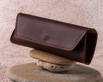 May include: A brown leather glasses case with a stitched edge and a small logo on the front. The case is resting on a stack of rocks.