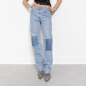May include: A woman wearing a black satin top with lace trim and light blue jeans with patches. She is standing with her hands on her hips and looking up.