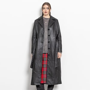 May include: A long black leather coat featuring a notched collar, a front button closure, and a matching tie belt. The coat is worn over a grey long-sleeve top, a red and black plaid skirt, and dark grey pointed-toe boots with black accents.