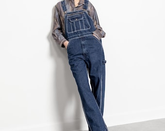 May include: Dark blue denim overalls featuring a front bib pocket with a button closure, side pockets, and straight-leg trousers. The overalls are worn over a long-sleeved, button-down shirt with a muted, abstract brown and gray pattern.