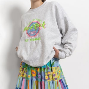 May include: A light gray crewneck sweatshirt featuring a Hard Rock Cafe St. Thomas logo with green and pink embroidery, paired with a tiered skirt showcasing a colorful patchwork pattern of floral and striped designs in blue, yellow, and pink.