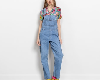 May include: Light blue denim overalls featuring a front bib pocket, side pockets, and adjustable shoulder straps, worn over a short-sleeved shirt with a vibrant multicolored floral pattern and bright yellow platform sandals.