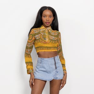 May include: A long-sleeved cropped shirt featuring a mustard yellow base with a multi-colored paisley and floral pattern, a pointed collar, and a smocked waist, paired with a light blue denim mini skirt featuring an off-center vertical zipper.
