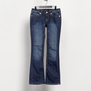 May include: Dark wash denim jeans with a slight flare at the hem, hanging from a black hanger. The jeans feature gold-colored rivets and a button closure. White stitching accents the pockets and seams. The jeans are displayed against a plain white background.