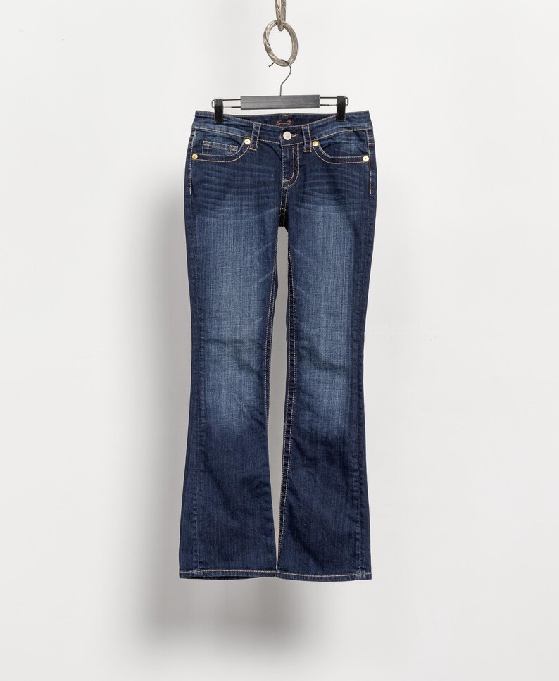 May include: Dark wash denim jeans with a slight flare at the hem, hanging from a black hanger. The jeans feature gold-colored rivets and a button closure. White stitching accents the pockets and seams. The jeans are displayed against a plain white background.