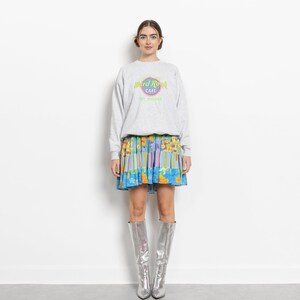 May include: A light gray crewneck sweatshirt featuring a Hard Rock Cafe St. Thomas logo in neon green and pink, paired with a multi-colored floral and striped tiered mini skirt and knee-high metallic silver pointed-toe boots.