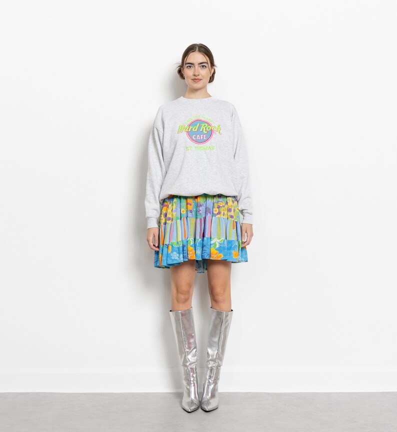 May include: A light gray crewneck sweatshirt featuring a Hard Rock Cafe St. Thomas logo in neon green and pink, paired with a multi-colored floral and striped tiered mini skirt and knee-high metallic silver pointed-toe boots.