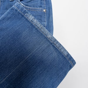 Pode incluir: Close-up de cal&ccedil;as de ganga azuis. As cal&ccedil;as de ganga t&ecirc;m um design cl&aacute;ssico de cinco bolsos com rebites prateados e fecho de bot&atilde;o. A bainha est&aacute; dobrada e as costuras s&atilde;o vis&iacute;veis. As cal&ccedil;as de ganga est&atilde;o penduradas num cabide preto sobre um fundo branco.