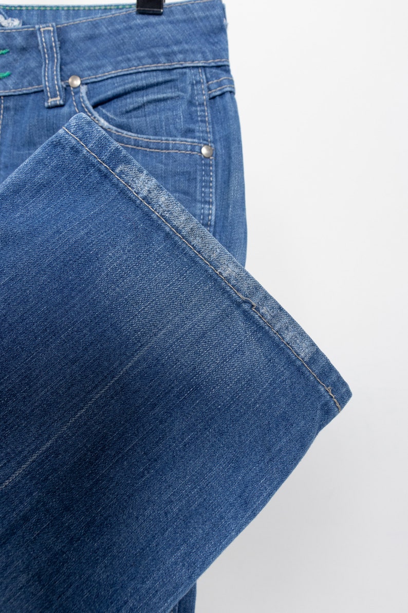 Pode incluir: Close-up de cal&ccedil;as de ganga azuis. As cal&ccedil;as de ganga t&ecirc;m um design cl&aacute;ssico de cinco bolsos com rebites prateados e fecho de bot&atilde;o. A bainha est&aacute; dobrada e as costuras s&atilde;o vis&iacute;veis. As cal&ccedil;as de ganga est&atilde;o penduradas num cabide preto sobre um fundo branco.