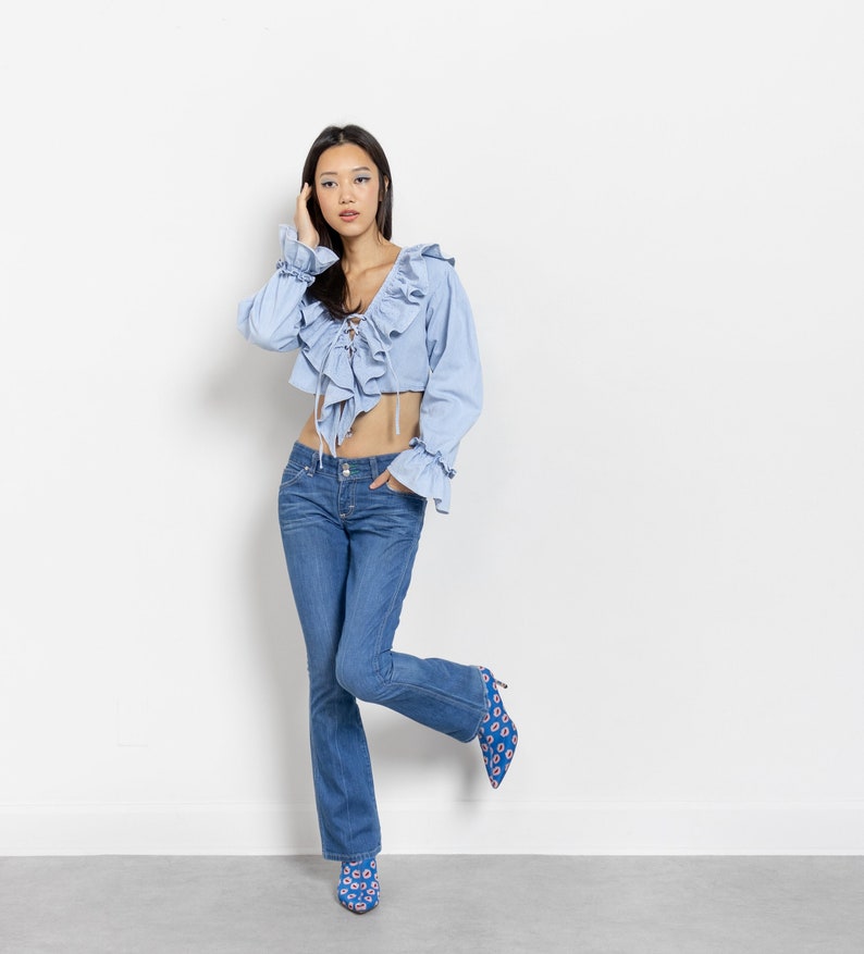 Pode incluir: Um top cropped azul claro com detalhes de babados e frente com cadar&ccedil;o &eacute; combinado com jeans azuis. O traje &eacute; complementado com saltos azuis estampados. A modelo est&aacute; em frente a uma parede branca.