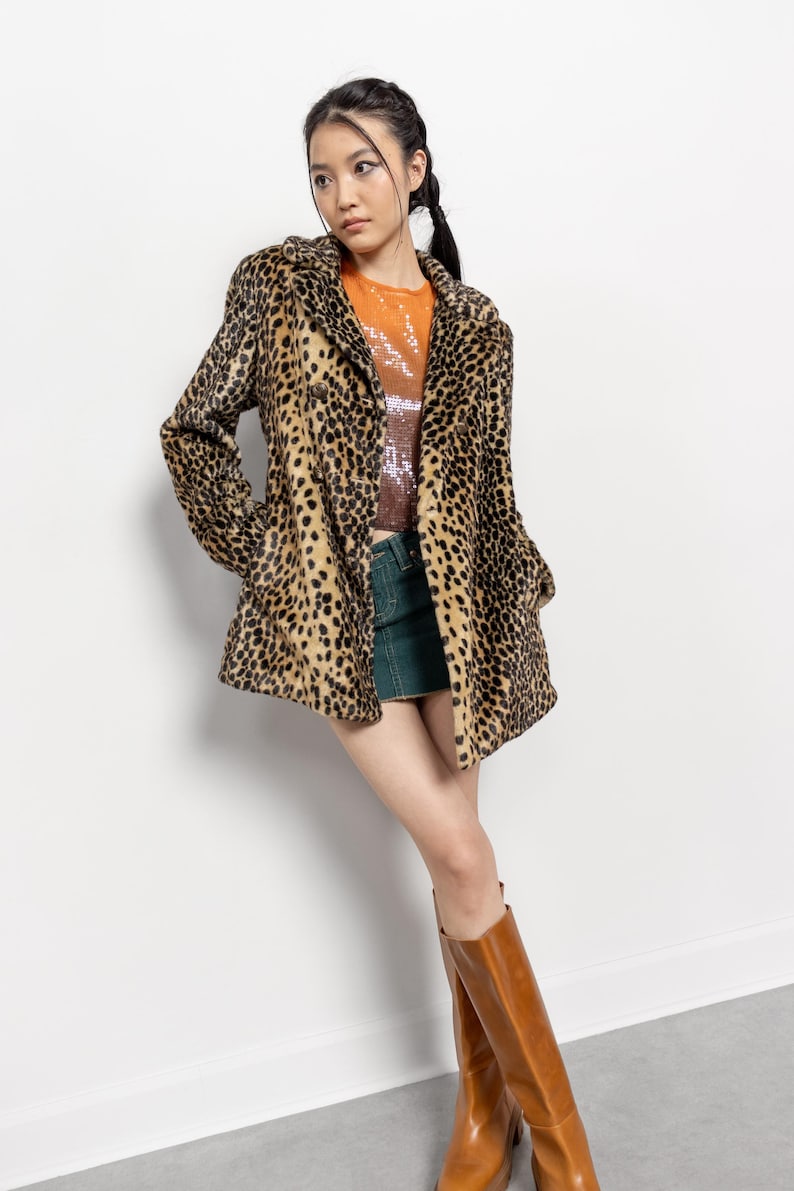 May include: A leopard print coat with a collared neckline and button closure. The coat is paired with a sequined top, a green denim skirt, and brown knee-high boots. The outfit is styled against a white background.