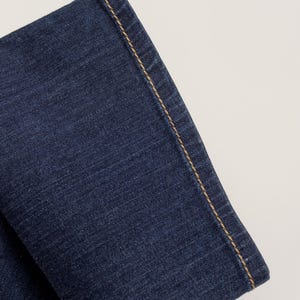 May include: Close-up of a section of dark blue denim jeans. The edge of the fabric is visible, with a contrasting tan-colored stitch running along the seam. The denim has a textured appearance.