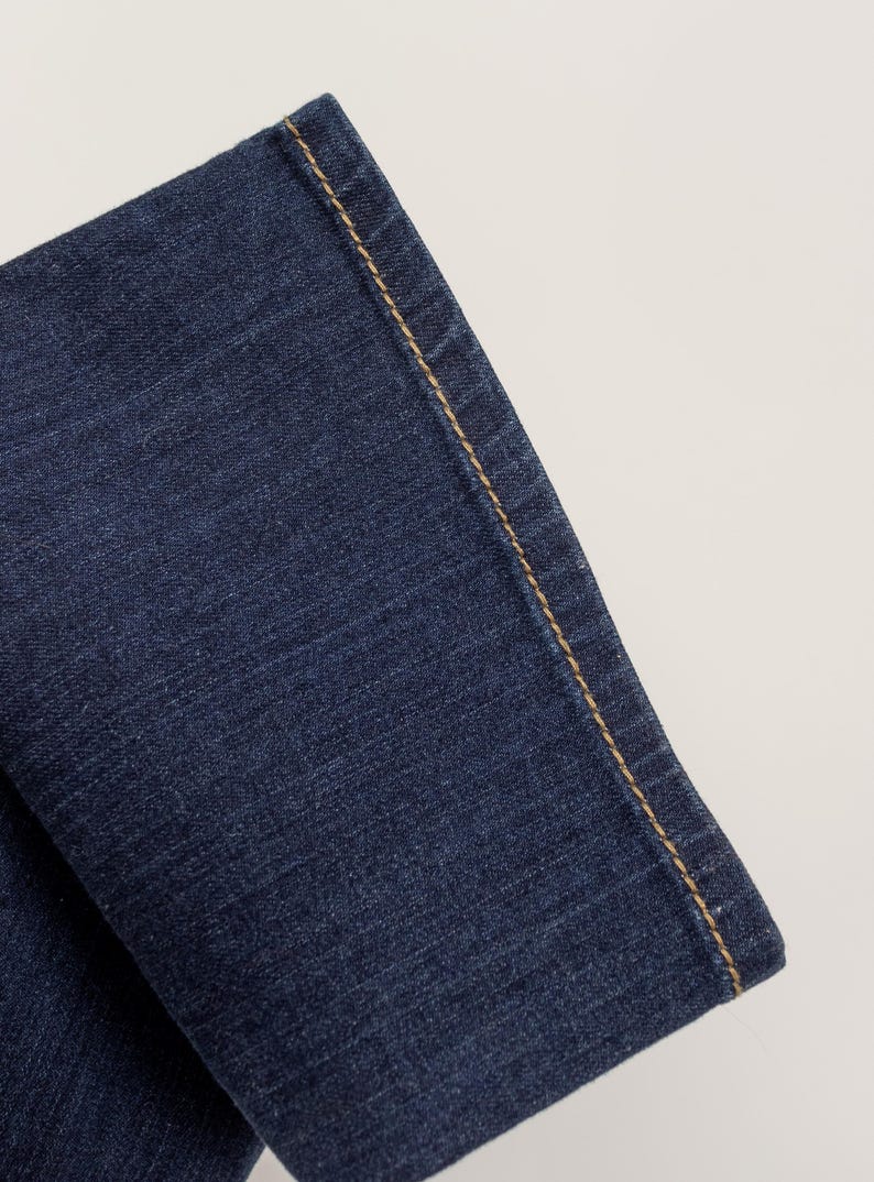 May include: Close-up of a section of dark blue denim jeans. The edge of the fabric is visible, with a contrasting tan-colored stitch running along the seam. The denim has a textured appearance.