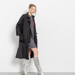 Puede incluir: Una mujer con un abrigo largo negro sobre un vestido gris, combinados con botas altas plateadas. Las botas tienen punta y tac&oacute;n cuadrado. El conjunto se presenta sobre un fondo blanco.