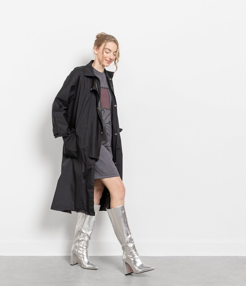 Puede incluir: Una mujer con un abrigo largo negro sobre un vestido gris, combinados con botas altas plateadas. Las botas tienen punta y tac&oacute;n cuadrado. El conjunto se presenta sobre un fondo blanco.