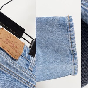May include: Light blue denim jeans featuring a tan leather patch on the back waistband, a small red fabric tag on the back pocket, reinforced stitching along the seams, and a straight-leg hem with visible fading and distressed texture details.