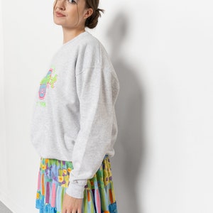 May include: A light gray long-sleeve sweatshirt featuring a Hard Rock Cafe logo in neon green, pink, and blue on the chest, paired with a tiered mini skirt showcasing a vibrant multicolored floral and striped pattern in shades of blue, yellow, and pink.