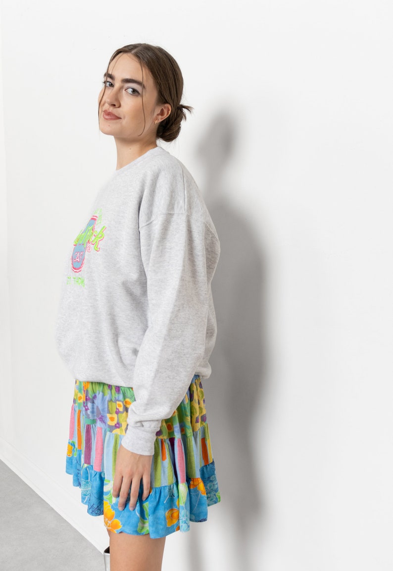 May include: A light gray long-sleeve sweatshirt featuring a Hard Rock Cafe logo in neon green, pink, and blue on the chest, paired with a tiered mini skirt showcasing a vibrant multicolored floral and striped pattern in shades of blue, yellow, and pink.