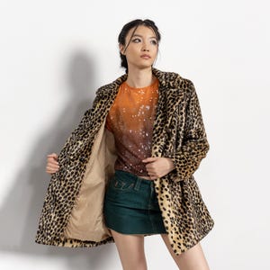 May include: A leopard print coat with a tan lining. The coat is open, revealing an orange and brown sequined top and a green denim skirt. The coat has a classic lapel and button closure.