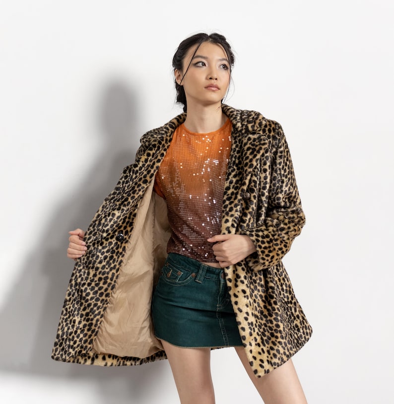 May include: A leopard print coat with a tan lining. The coat is open, revealing an orange and brown sequined top and a green denim skirt. The coat has a classic lapel and button closure.