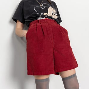 Puede incluir: Una camiseta negra con un gráfico de Mickey Mouse y pantalones cortos de pana rojos. Los pantalones cortos tienen cintura alta y pliegues. La persona también lleva calcetines altos grises.