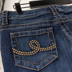 May include: Close-up of blue denim jeans with a decorative pocket. The pocket features a gold-colored stitched design. The jeans are hanging on a black hanger.