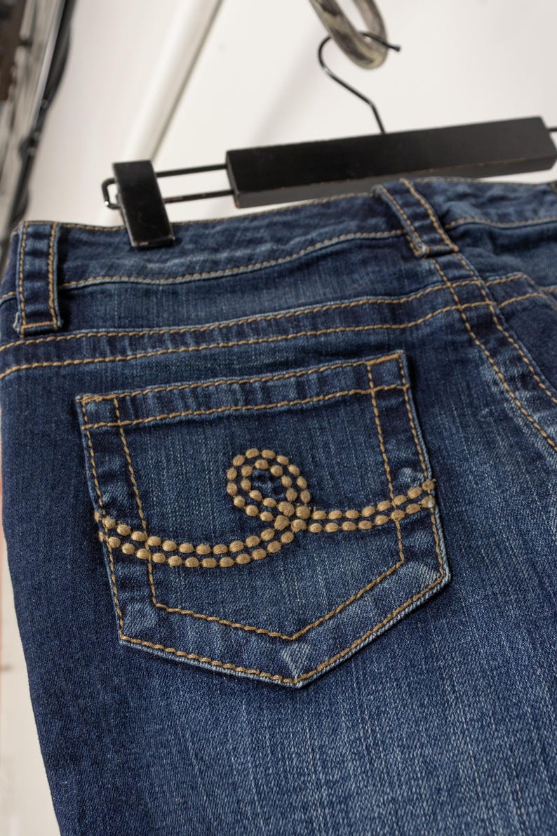 May include: Close-up of blue denim jeans with a decorative pocket. The pocket features a gold-colored stitched design. The jeans are hanging on a black hanger.