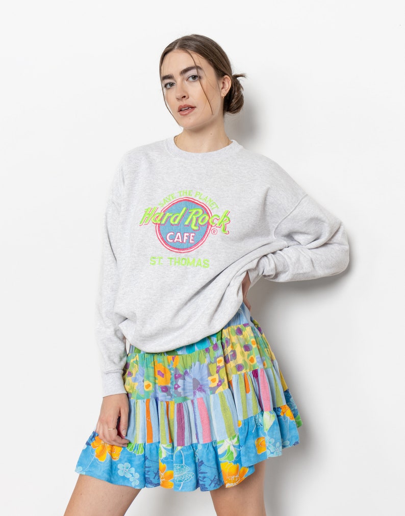 May include: A light gray crewneck sweatshirt featuring a Hard Rock Cafe St. Thomas logo with neon green and pink text. Paired with a tiered mini skirt displaying a vibrant patchwork pattern of blue, yellow, and pink floral and striped prints.