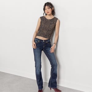 May include: A woman wearing a sleeveless, textured brown top and dark wash blue jeans with a slight flare. The jeans have decorative stitching. She is also wearing maroon pointed-toe heels.