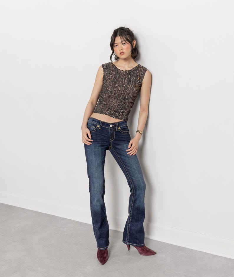 May include: A woman wearing a sleeveless, textured brown top and dark wash blue jeans with a slight flare. The jeans have decorative stitching. She is also wearing maroon pointed-toe heels.