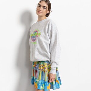 May include: A light gray crewneck sweatshirt featuring a Hard Rock Cafe St. Thomas logo in neon green and pink on the chest, paired with a tiered mini skirt showcasing a vibrant patchwork pattern of blue, yellow, and orange floral and striped prints.