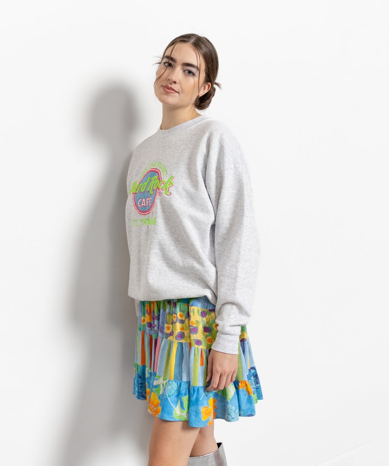 May include: A light gray crewneck sweatshirt featuring a Hard Rock Cafe St. Thomas logo in neon green and pink on the chest, paired with a tiered mini skirt showcasing a vibrant patchwork pattern of blue, yellow, and orange floral and striped prints.