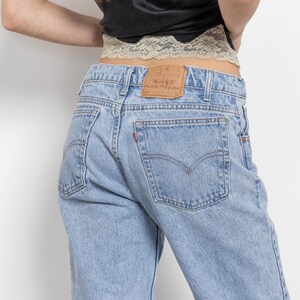 May include: Light blue denim jeans featuring a tan leather patch on the back waistband, two back pockets with stitching details, and a small red tab on the right pocket. The jeans are worn with a black top and a beige lace trim detail at the waist.