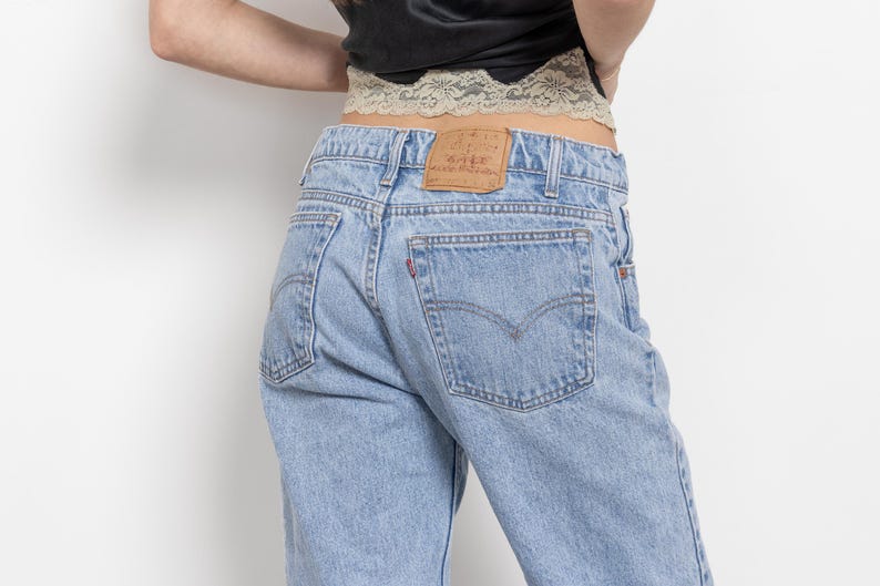 May include: Light blue denim jeans featuring a tan leather patch on the back waistband, two back pockets with stitching details, and a small red tab on the right pocket. The jeans are worn with a black top and a beige lace trim detail at the waist.