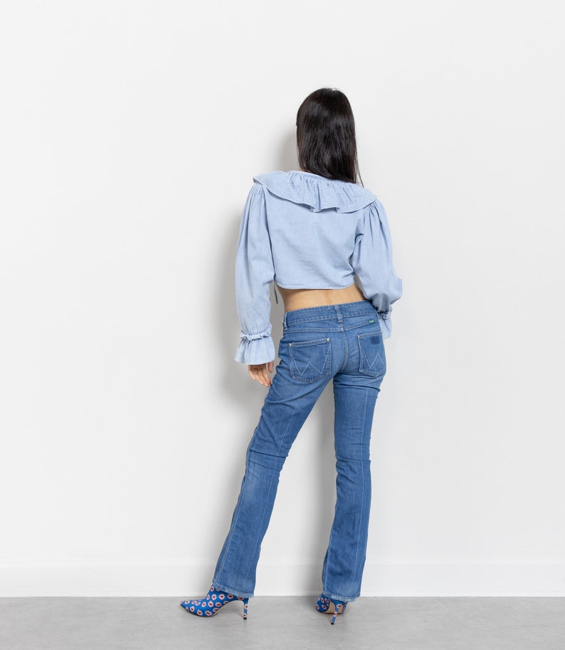 Pode incluir: Blusa cropped azul claro com gola com babados e mangas compridas, combinada com jeans azuis. Os jeans t&ecirc;m pernas largas e s&atilde;o usados com sapatos de salto alto estampados. O conjunto &eacute; apresentado sobre um fundo branco.