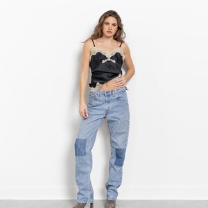 May include: A black satin camisole featuring beige lace trim along the neckline and hem, paired with light blue denim jeans that have darker blue rectangular patches on the knees and gray patterned pointed-toe ankle boots.