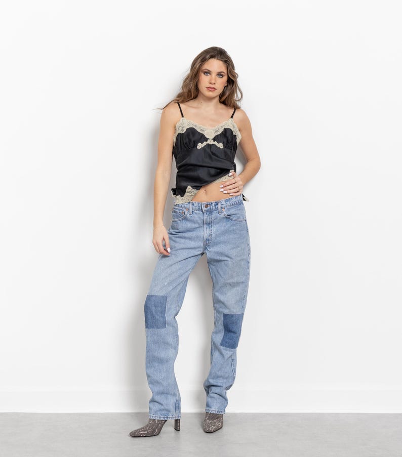 May include: A black satin camisole featuring beige lace trim along the neckline and hem, paired with light blue denim jeans that have darker blue rectangular patches on the knees and gray patterned pointed-toe ankle boots.