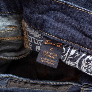 May include: Close-up of dark blue denim jeans. The tag reads "Seven7" with size 4, 99% cotton, and 1% spandex. The jeans have a classic design with a leather patch and decorative stitching.
