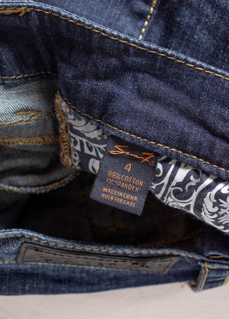 May include: Close-up of dark blue denim jeans. The tag reads "Seven7" with size 4, 99% cotton, and 1% spandex. The jeans have a classic design with a leather patch and decorative stitching.