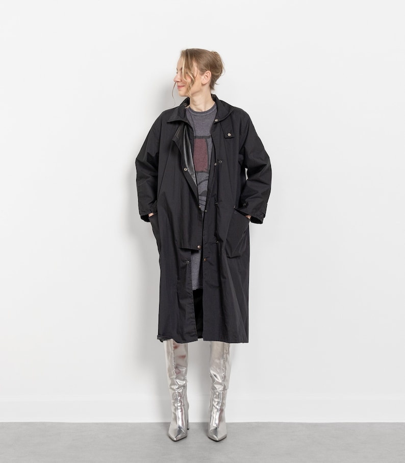 Puede incluir: Un abrigo largo negro con cierre de botones y bolsillos. El abrigo se lleva sobre una camiseta gris y un vestido gris. Botas plateadas hasta la rodilla con punta completan el conjunto. El abrigo es una prenda de vestir exterior cl&aacute;sica.