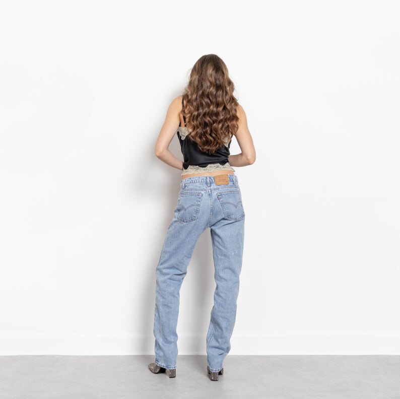 May include: A black camisole with beige lace trim along the neckline and hem paired with light blue denim jeans. The jeans feature a classic five-pocket design, a tan leather patch on the back waistband, and a straight-leg cut with a medium wash.